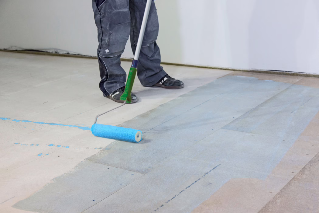 UZIN_Application Image_PE360 Plus_Renoplan System_2020-10 Kuvan alaosassa näkyvät työtekijän sääret ja jalat, kun hän levittää pitkällä maaliharjalla vaaleansinistä primeria tai maalia puulattian alustaan. Henkilöllä on tummat työhousut ja mustat lenkkarit; tela jättää lattialautojen pinnalle kostean sinisen kerroksen valkoseinämisessä sisätilassa. Kuva korostaa lattiavalmistelua, työkaluja ja maalitahroja.
